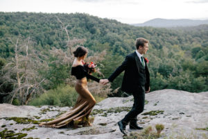 Bride, wearing a black shirt and gold skirt, holding hands with groom, wearing a black suit and green tie, walking on a mountain top at Mill Creek Farm in Hiddenite NC.