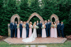 Bride, in white dress, and Groom, in blue suit, with wedding party laughing in background atRolling Hill Farm Wedding Venue in Monroe NC.