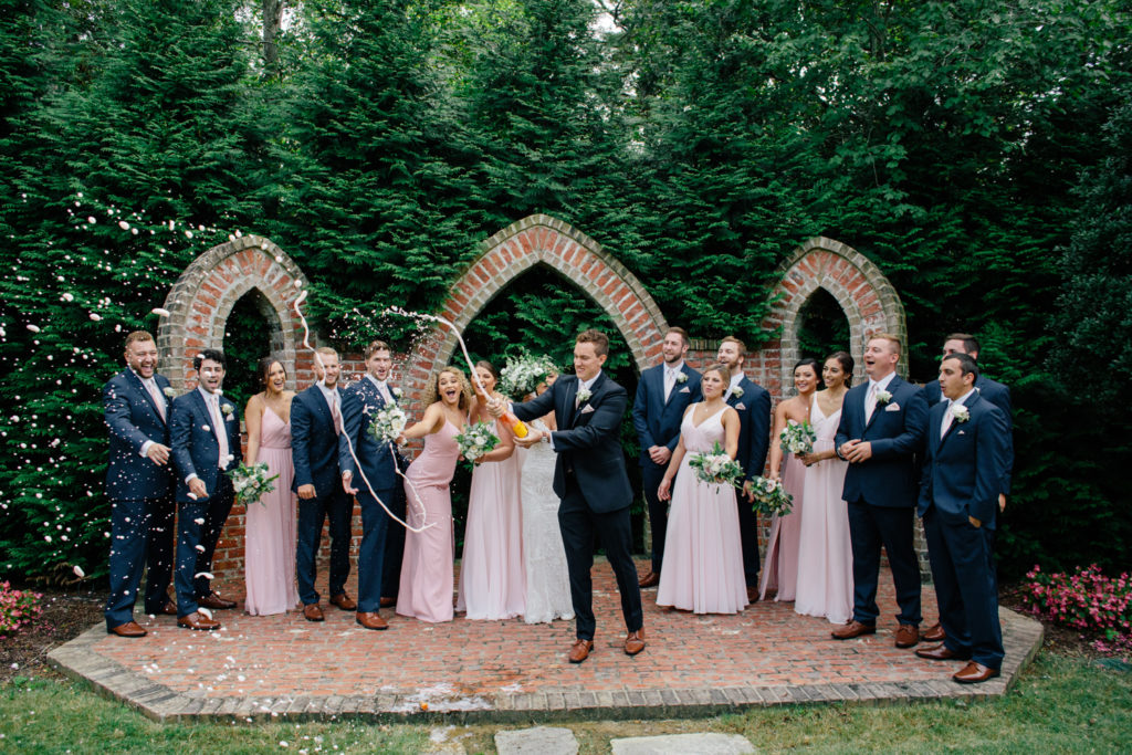 Groom popping champagne bottle with wedding party laughing in background atRolling Hill Farm Wedding Venue in Monroe NC.