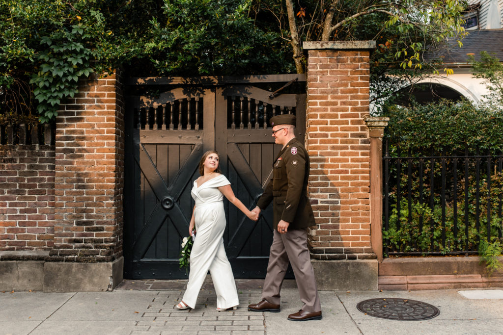 Bride, in white jumpsuit, walking with her groom, in green military uniform, at the White Point Gardens in Charleston.