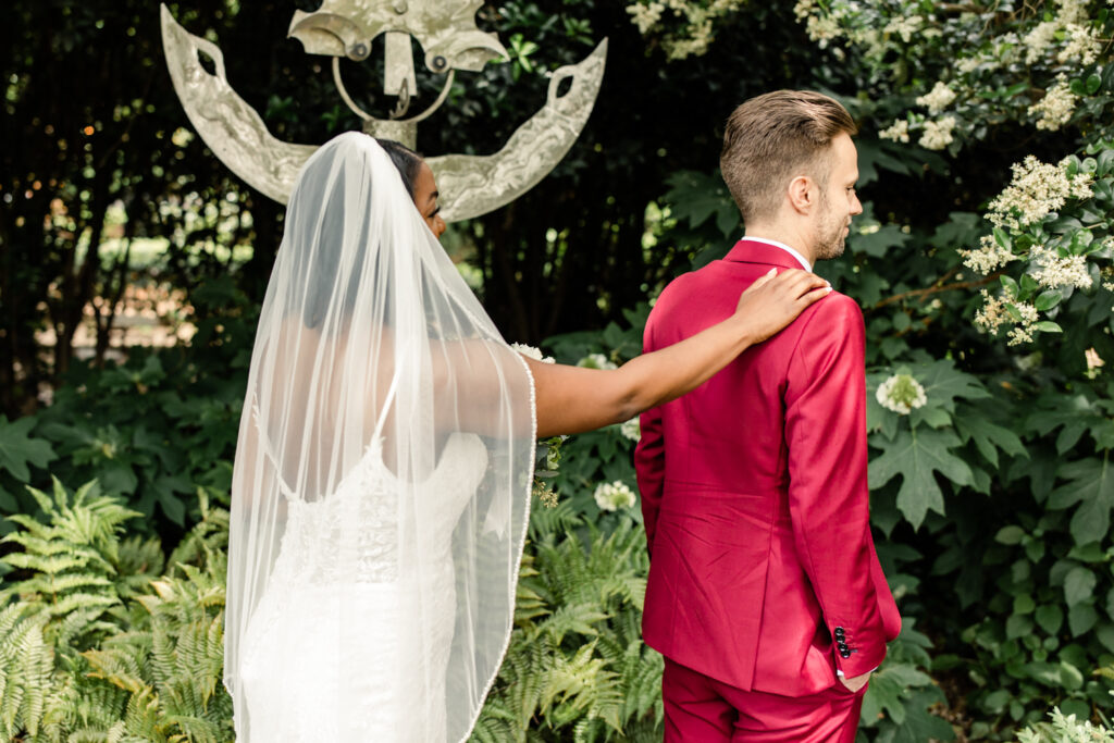 Bride, in white gown, touching the right shoulder of groom, in red suit, during first touch at McGill Rose Garden in Charlotte NC.