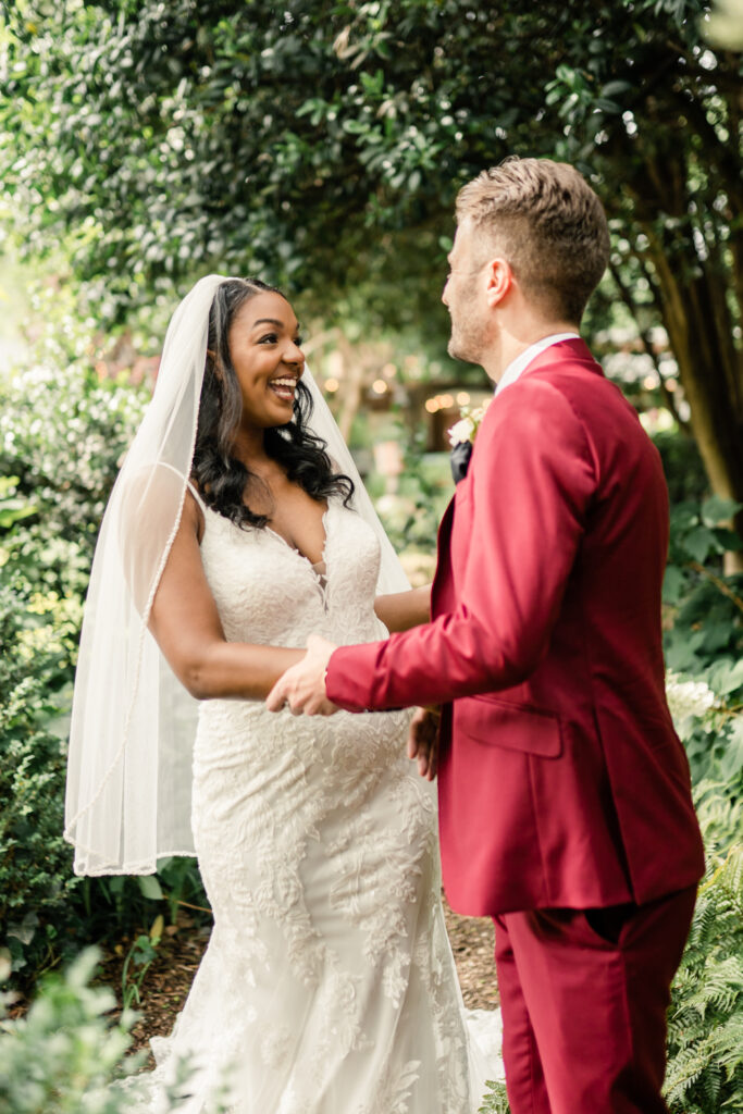 Bride, in white gown, laughing with her groom, in red suit, during first touch at McGill Rose Garden in Charlotte NC.