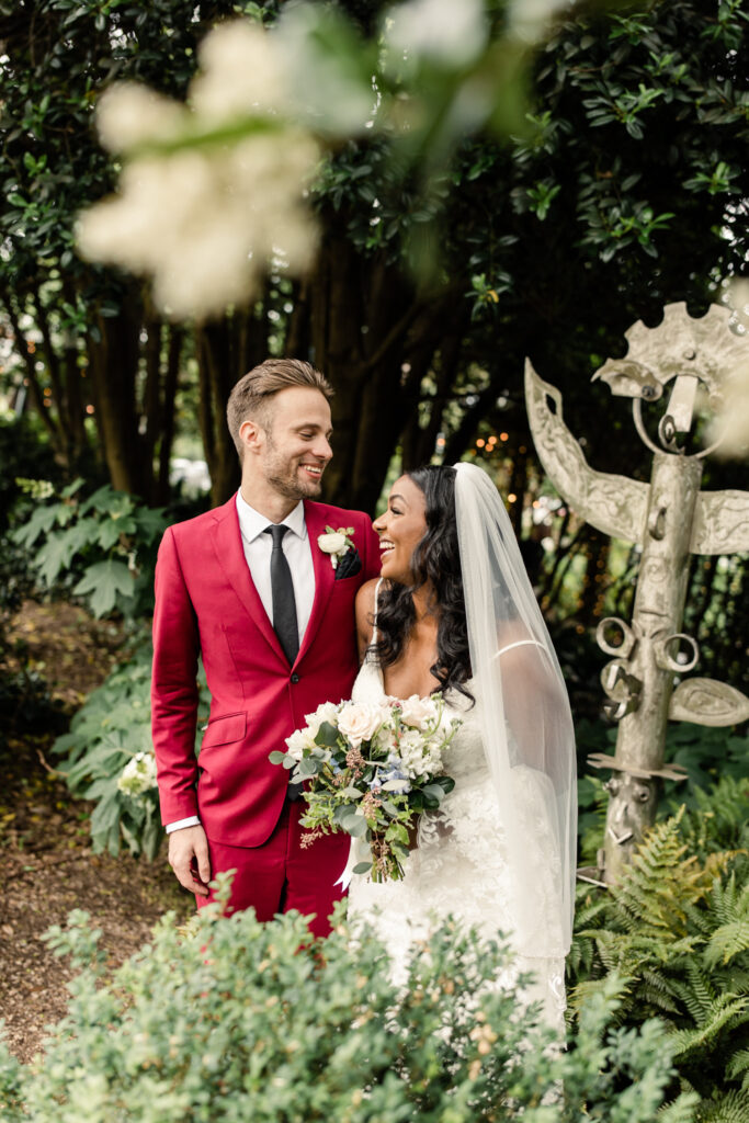 Bride, in white gown holding white flowers, laughing with her groom, in red suit, during first touch at McGill Rose Garden in Charlotte NC.