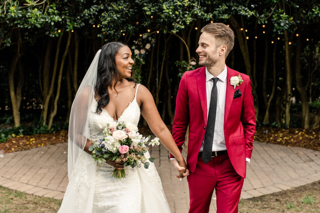 Bride, in white gown, walking holding hands with groom, in red suit, at McGill Rose Garden in Charlotte NC white greenery and twinkly lights in background.