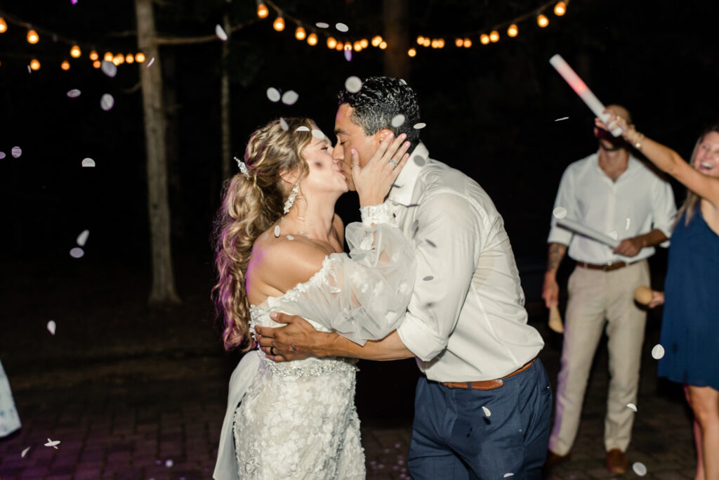 Bride, in white lace dress, kisses groom, in white button down and blue pants, under confetti at night during reception exit at Morning Glory Farm Wedding Venue in Charlotte NC.