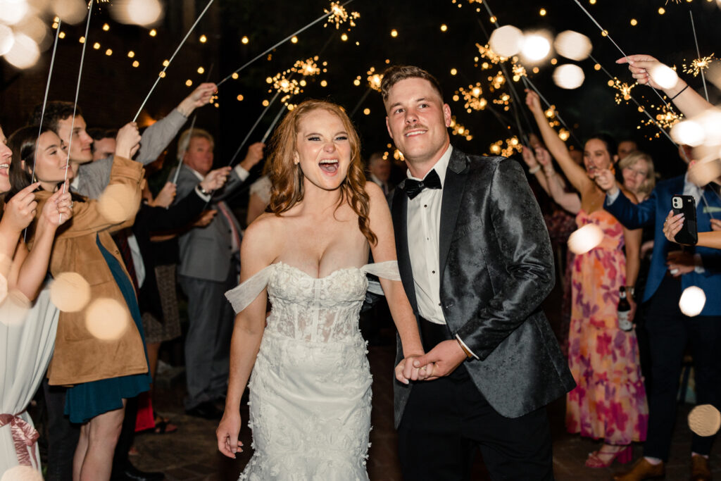 Bride, in white lace dress, holding hands with groom, in black suit, during sparkler exit at The Millstone at Adams Pond Wedding Venue in Colombia SC