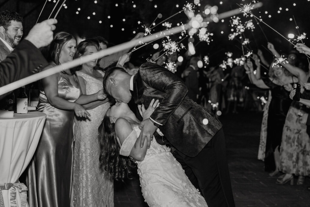 Bride, in white lace dress, holding hands and kissing groom, in black suit, during sparkler exit at The Millstone at Adams Pond Wedding Venue in Colombia SC