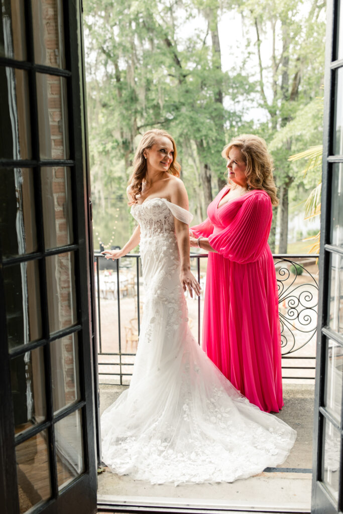 Bride, in white lace dress, getting into her dress with the help of her mom, in pink dress, zipping her up at The Millstone at Adams Pond Wedding Venue in Colombia SC