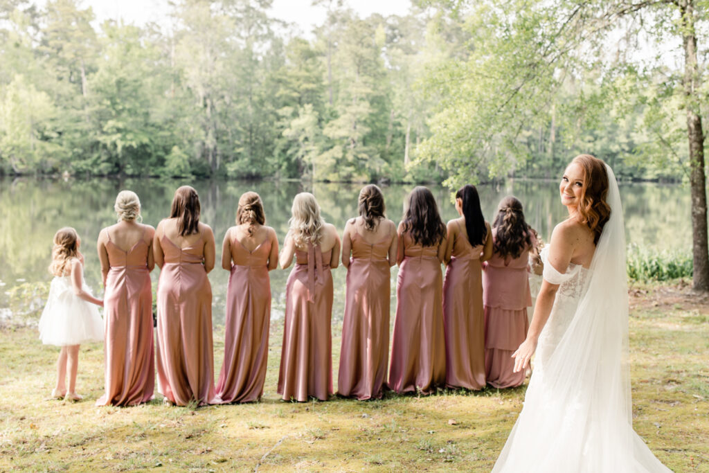 Eight bridesmaids, in pink dresses with pink bouquets, see bride, in white dress and veil, for the first time at The Millstone at Adams Pond Wedding Venue in Colombia SC