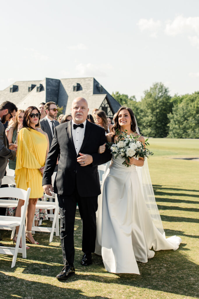 Bride, in white gown holding white bouquet, walking down aisle with father, in black suit, at Long View Country Club Wedding Venue Charlotte NC