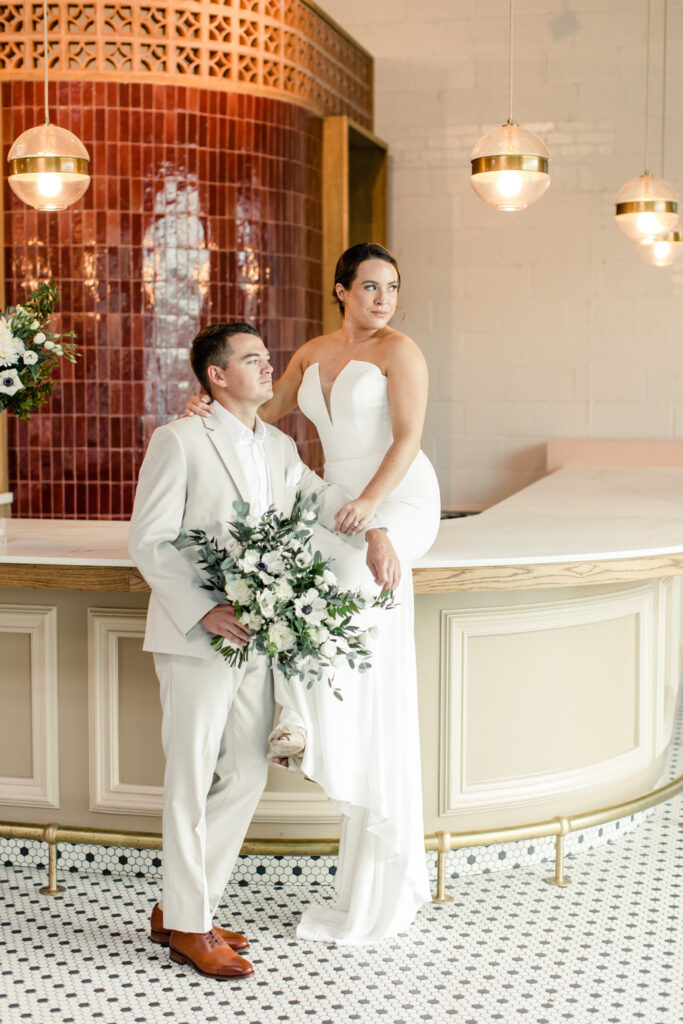 Bride, in white strapless dress, sitting on bar counter with arm around her groom, in a tan suit with white and green flowers at The Ruth wedding venue in Charlotte NC. Photographed by Charlotte Wedding Photographer.
