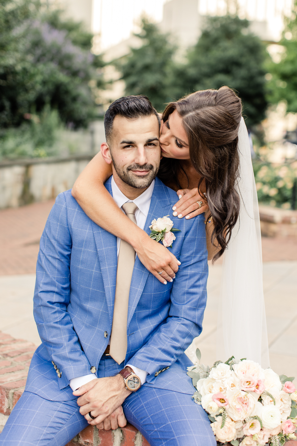 Groom, with brown hair, sitting on red brick wall with brunette bride kissing him on the cheek. Photographed by Charlotte wedding photographer