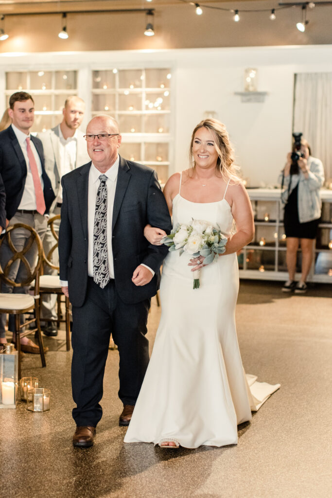 Bride with long blonde hair walking with father in black suit down the aisle at The Venue in Asheville NC during ceremony. Photographed by Charlotte wedding photographer.