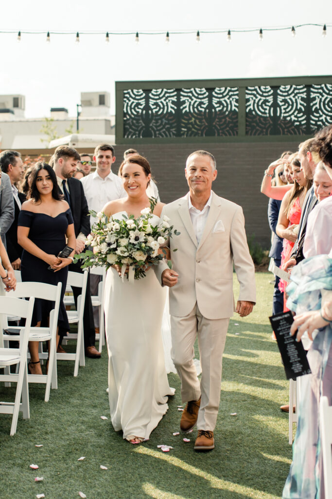 Bride, in white dress holding white and green floral bouquet, with her dad, in tan suit, at The Ruth wedding venue in Charlotte NC. Photographed by Charlotte Wedding Photographer.