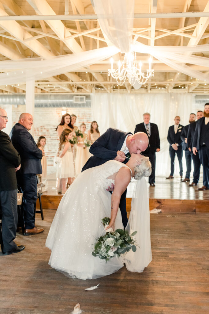 Bride with blonde hair in a white lace dress kissing groom, in a navy suit in the middle of ceremony aisle at The Bottle Factory Wedding Venue. Photographed by Charlotte wedding photographer.