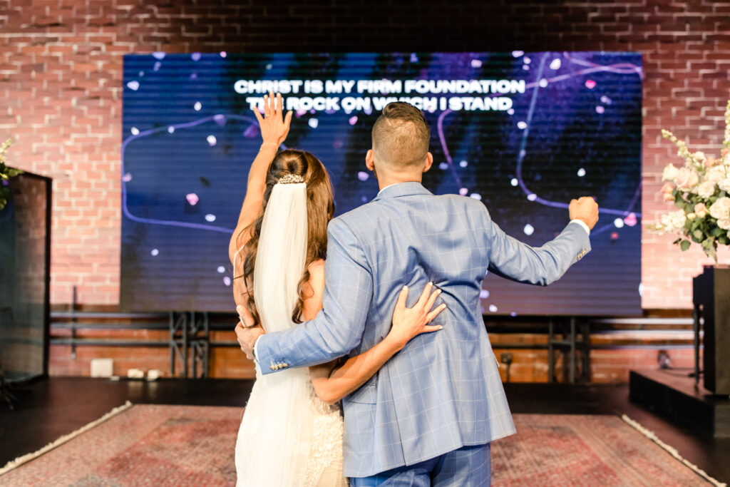 Bride in white dress and groom in blue suit praising God with hands in air photographed by Charlotte Wedding Photographer.  