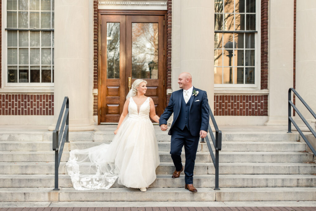 Bride with blonde hair in a white lace dress holding hands with groom, in a navy suit at The Bottle Factory Wedding Venue. Photographed by Charlotte wedding photographer.