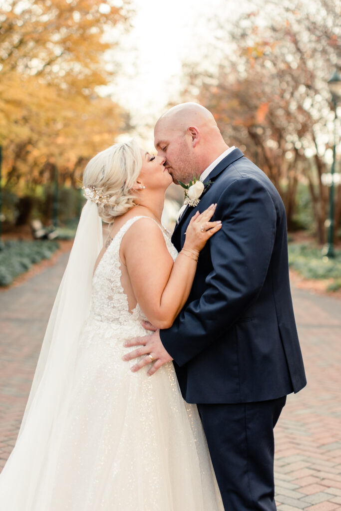 Bride with blonde hair in a white lace dress kissing groom, in a navy suit at The Bottle Factory Wedding Venue. Photographed by Charlotte wedding photographer.