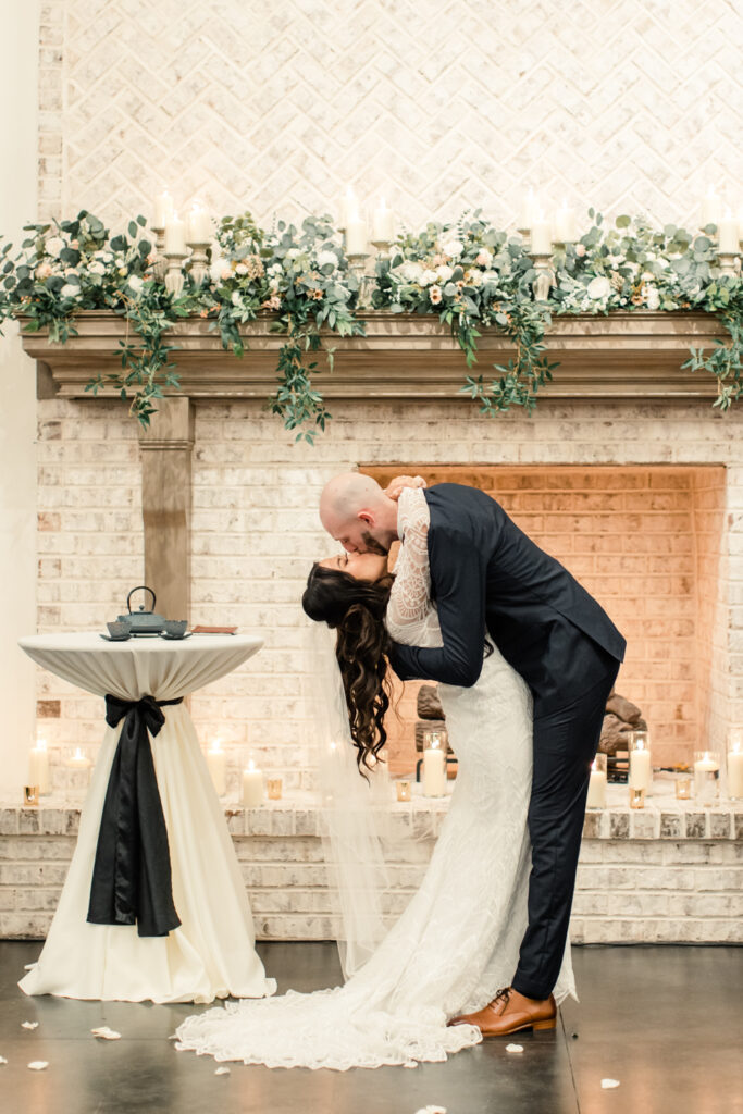 Bride in white long sleeve less dress and veil kissing her groom in blue suit during ceremony at Fields at Skycrest Wedding Venue in Charlotte NC. Photographed by Charlotte wedding photographer.