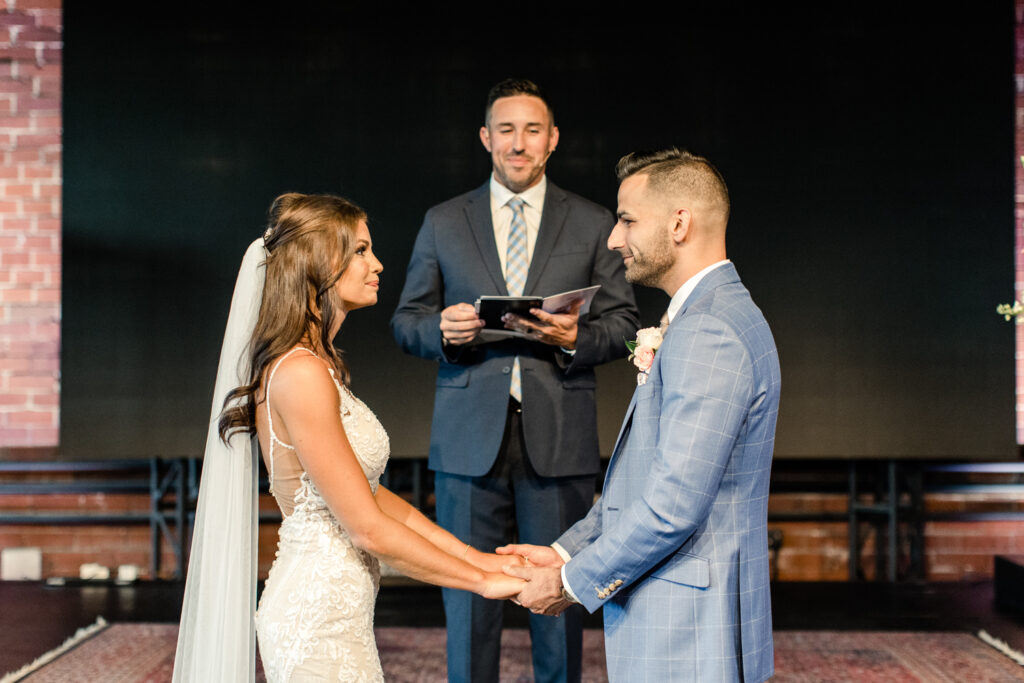 Bride with brown hair in white dress and veil holding hands with groom in blue suit and pink boutonniere in front of paster photographed by Charlotte Wedding Photographer.  
