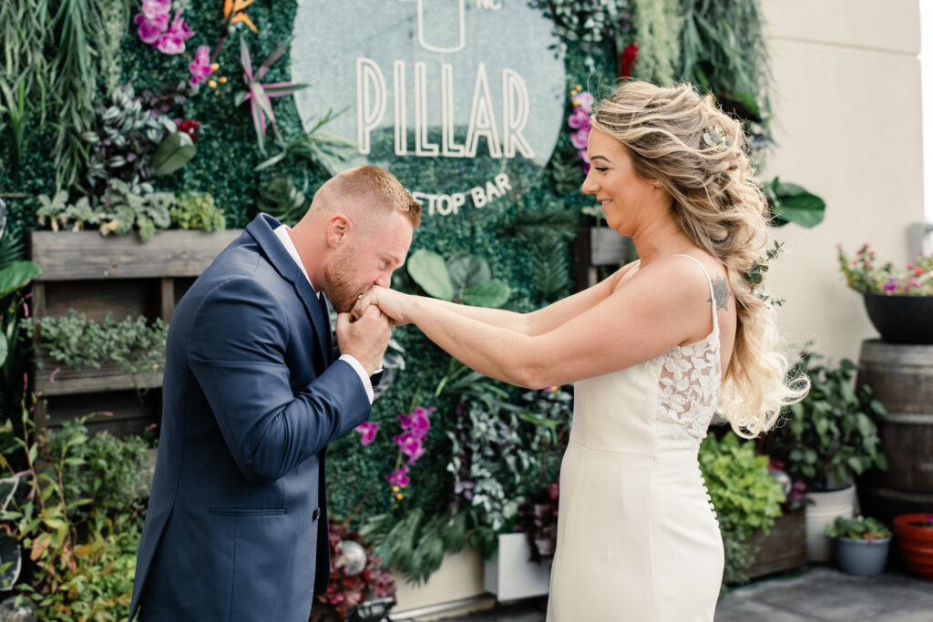 Groom with red hair wearing a blue suit kissing the hands of his bride who is in a white lace dress with long blonde hair. Photographed by Charlotte wedding photographer.